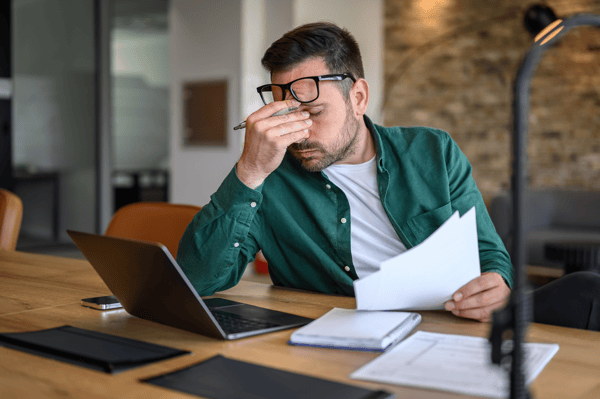 Mann sitzt im Büro mit Laptop und Dokumenten und hält sich gestresst den Kopf.